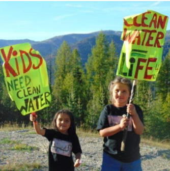 Two children stand outdoors holding signs reading "Kids Need Clean Water" and "Clean Water for Life," with a forested mountain backdrop. The mood is earnest.