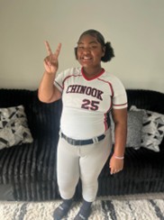 A middle school aged Black girl, Adenai, poses in front of a couch in jeans and a sports t-shirt and holding up a peace sign.