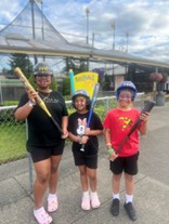 Three Black children wearing helmets and holding baseball bats pose outside.